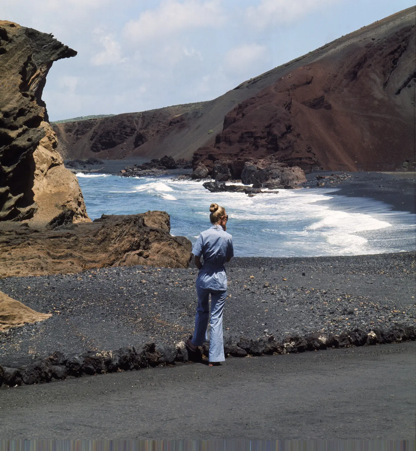 A woman on a black beach below red cliffs